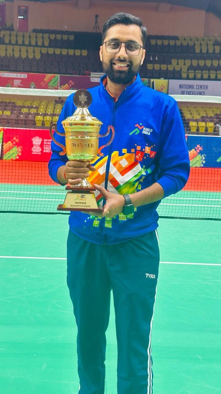 Suresh Kumar in a blue tracksuit holding the Khelo India University Games trophy on court